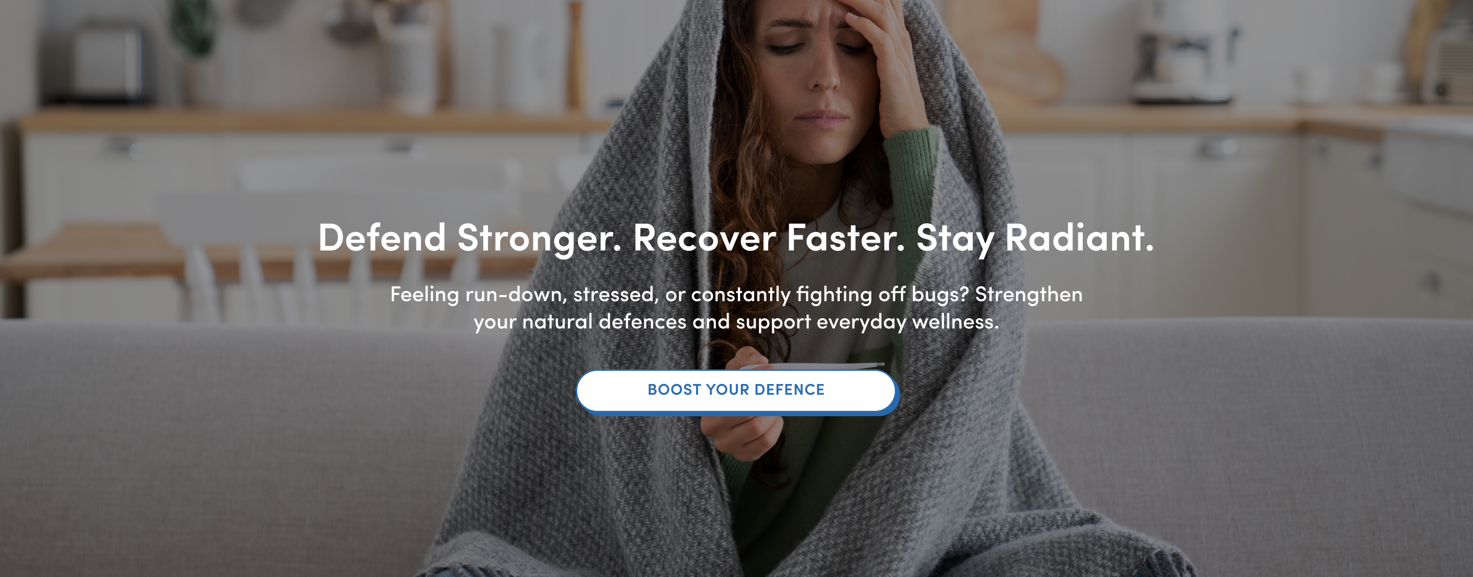 A woman sits on a couch wrapped in a gray blanket, looking unwell with her hand on her forehead. Text over the image promotes strengthening natural defenses and wellness, with a button labeled BOOST YOUR DEFENCE.