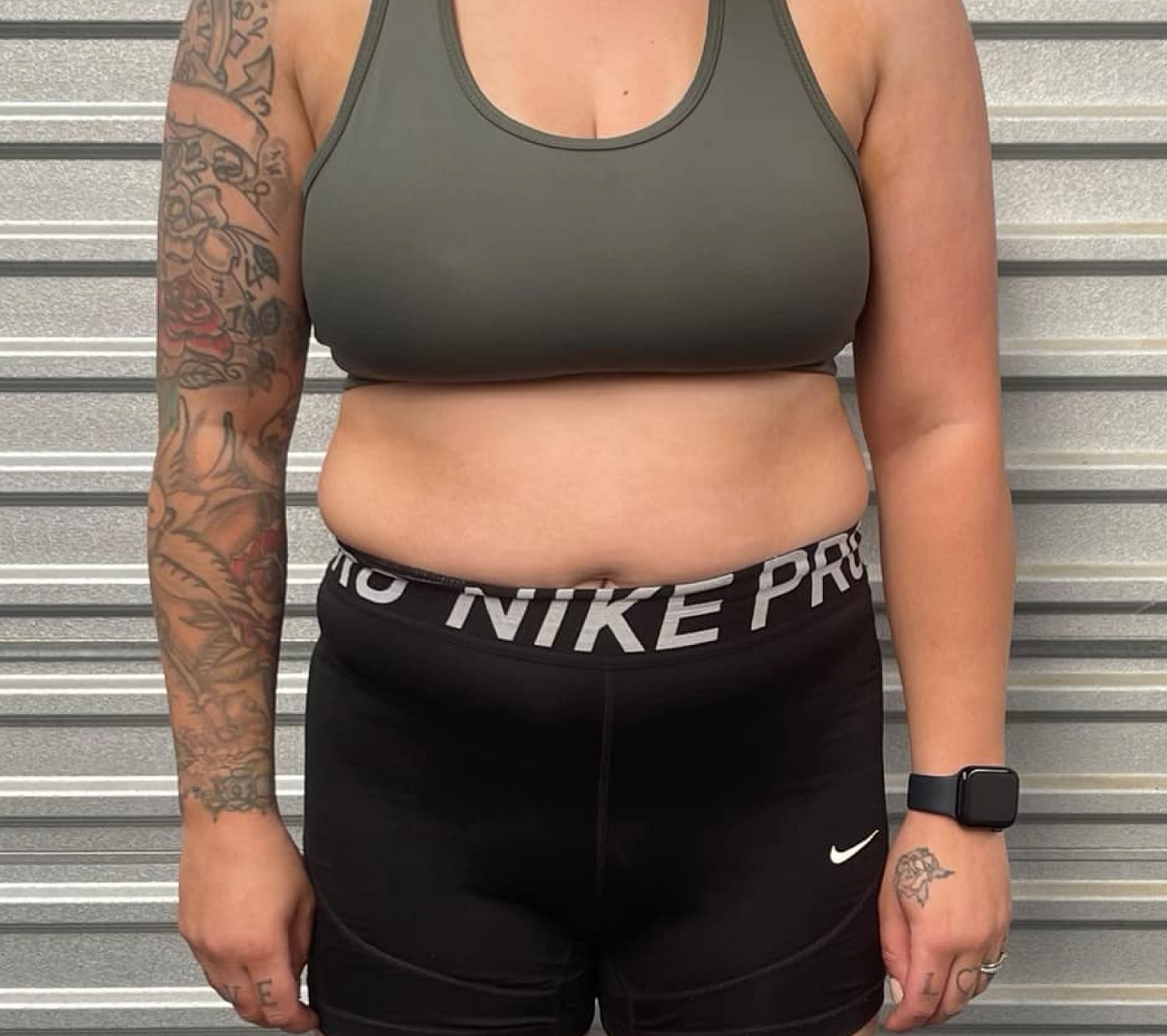 Person in workout attire with tattoos on arms stands against a metal background, wearing a dark sports bra and Nike Pro shorts.