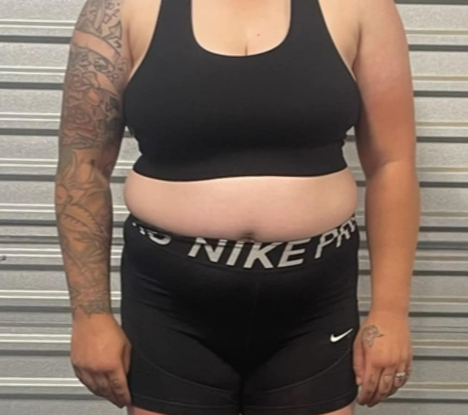 A person in a black sports top and shorts stands against a metal wall. One arm shows detailed tattoos.