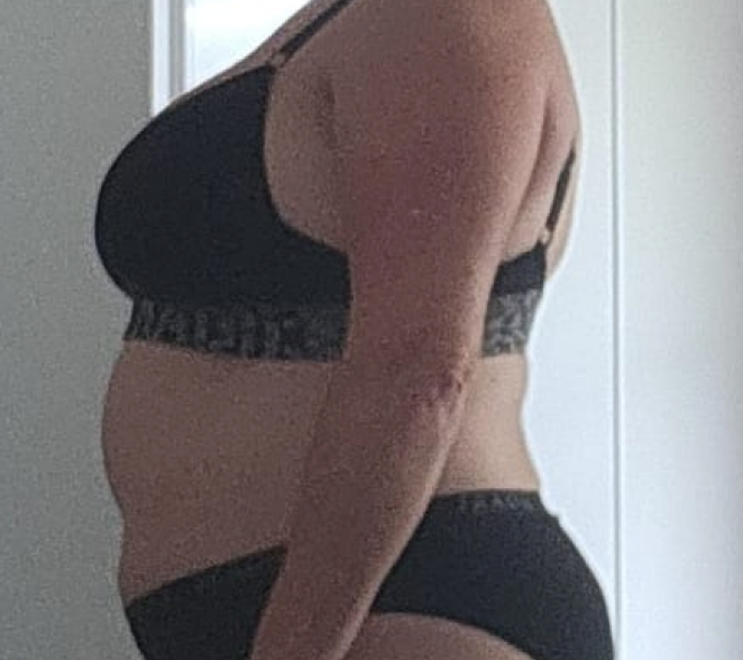 Side profile of a person in a black bra and underwear, standing in a room. The focus is on body shape and posture.
