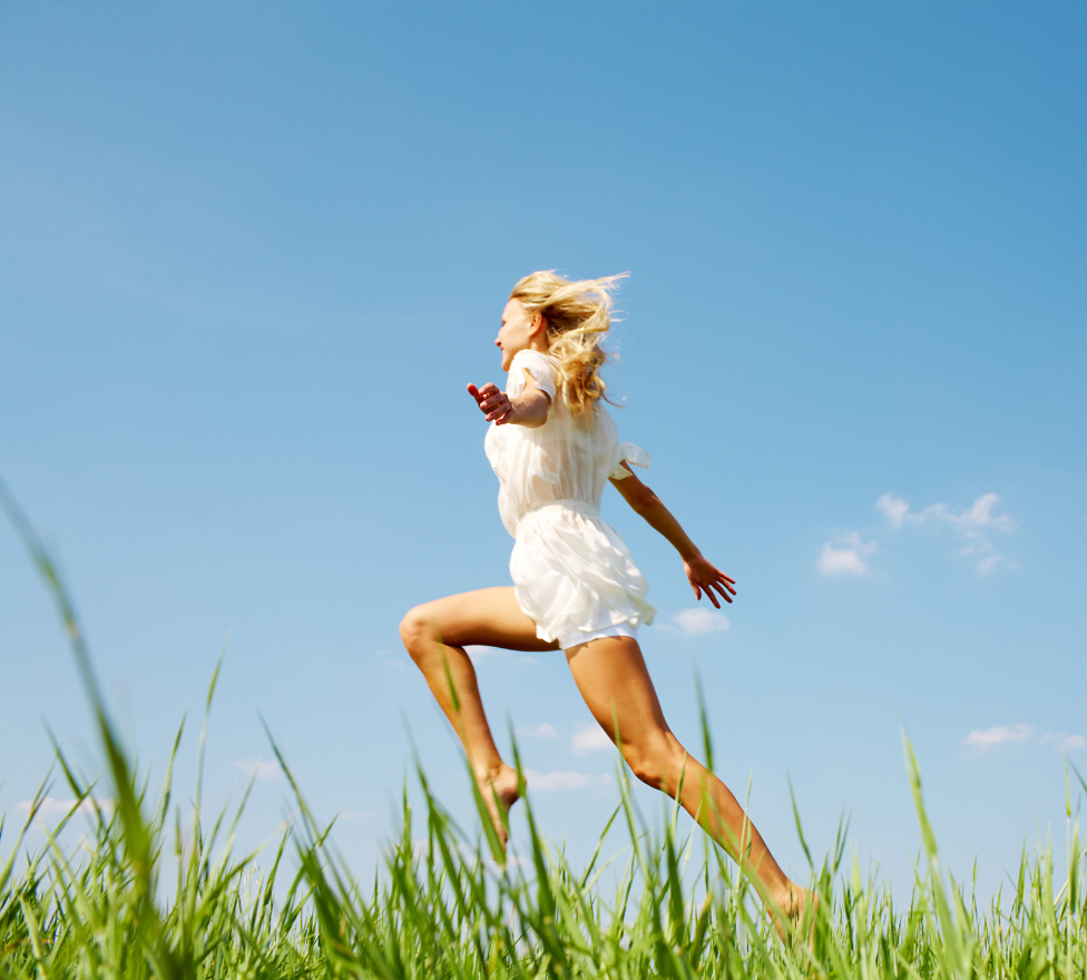 Woman in a white dress joyfully leaping through a sunny, grassy field under a clear blue sky.