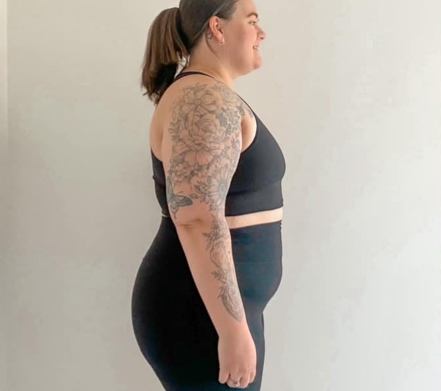 Profile of a person in black athletic wear with floral tattoo sleeve, standing against a plain background. Focus on fitness and body art.