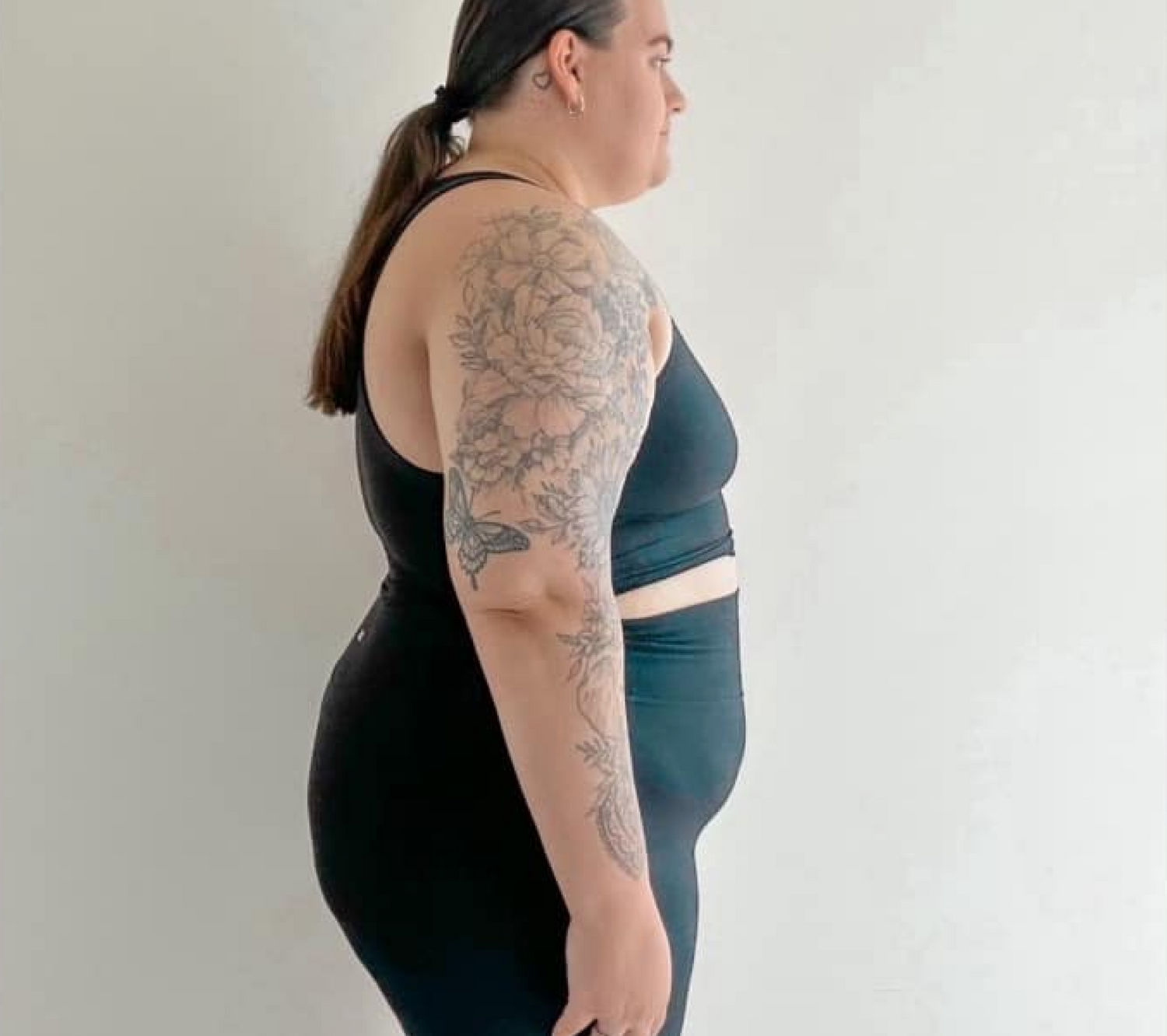 Person in black workout attire with floral arm tattoo standing side view. Clean background suggests focus on fitness and wellness.