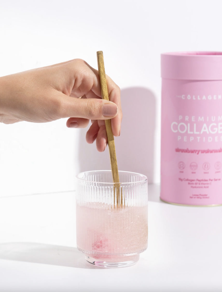 A hand stirs pink collagen drink in a glass with a wooden spoon; pink collagen container in the background.