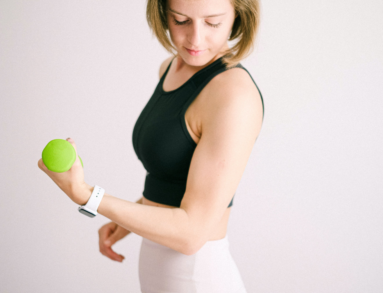 Woman in black sports top lifting a green dumbbell, wearing a white smartwatch, promoting fitness and beauty.