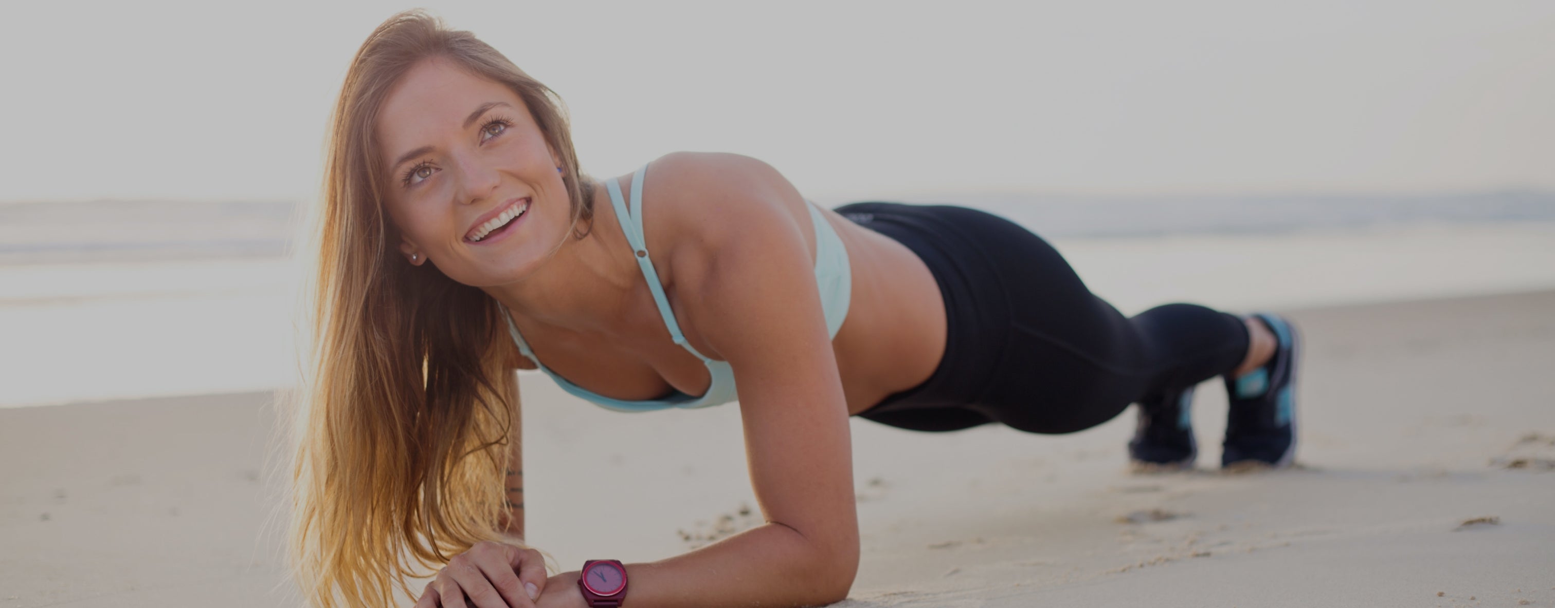 Smiling woman planking on a sunny beach, wearing a sports bra and leggings, embodying fitness and beauty lifestyle.
