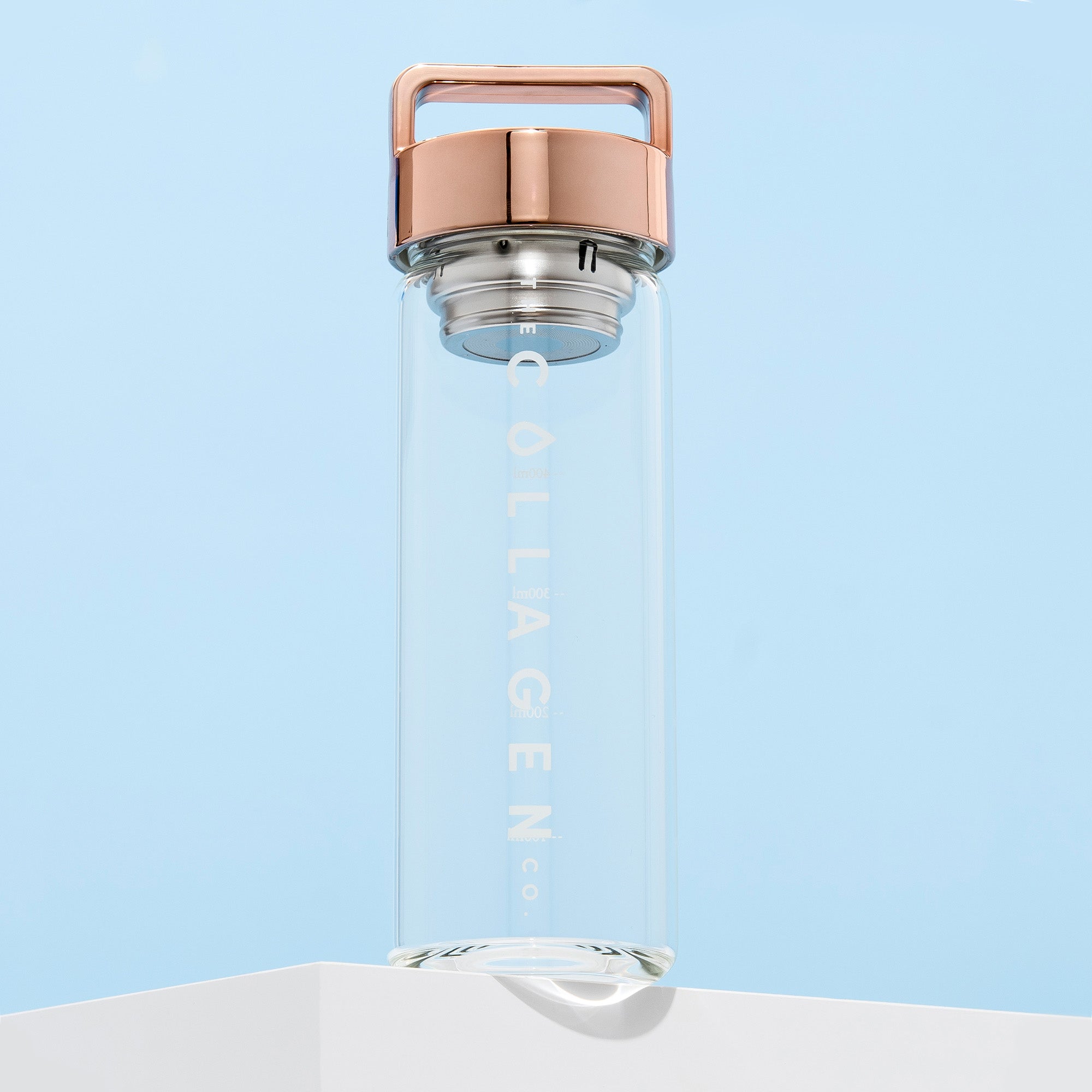 Clear glass bottle with "Collagen" text, rose gold lid on a white platform, set against a pale blue background. Ideal for beauty supplements.
