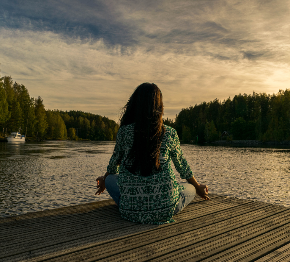 Woman meditating on dock by serene lake at sunset, surrounded by trees. Calm and peaceful setting, ideal for beauty and wellness reflection.