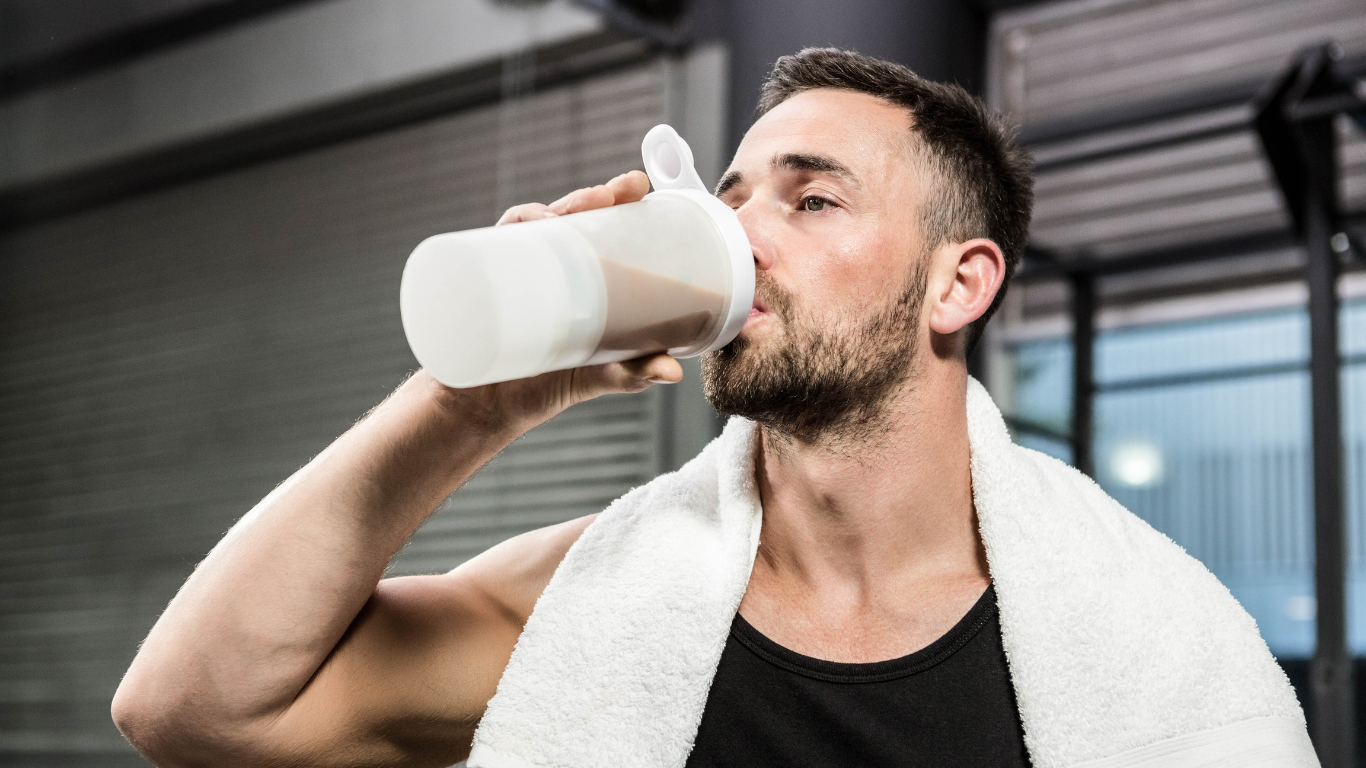 Man in gym with towel drinking from shaker, possibly collagen supplement. Enhancing beauty and wellness routine post workout.
