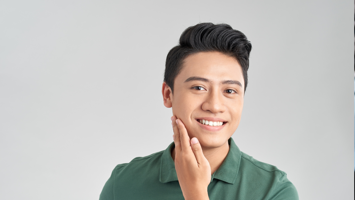 Man smiling, touching cheek, wearing green shirt. Smooth skin suggests collagen supplement use for beauty and skincare. Neutral background.