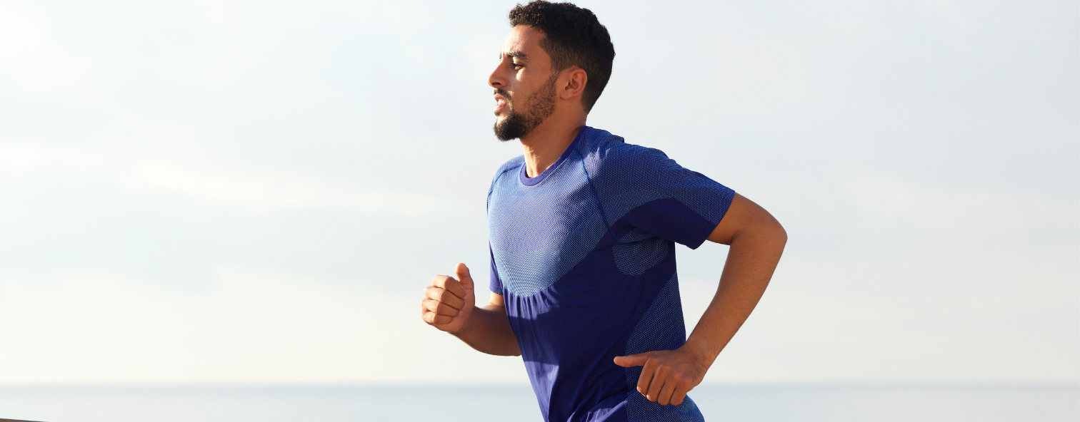 Man jogging outdoors by the sea in a blue shirt, showcasing fitness and wellness lifestyle.