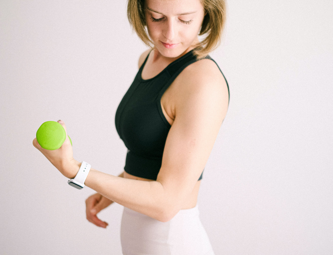 Woman in black sports top lifting a small green dumbbell, wearing a white smartwatch, focusing on fitness and wellness.