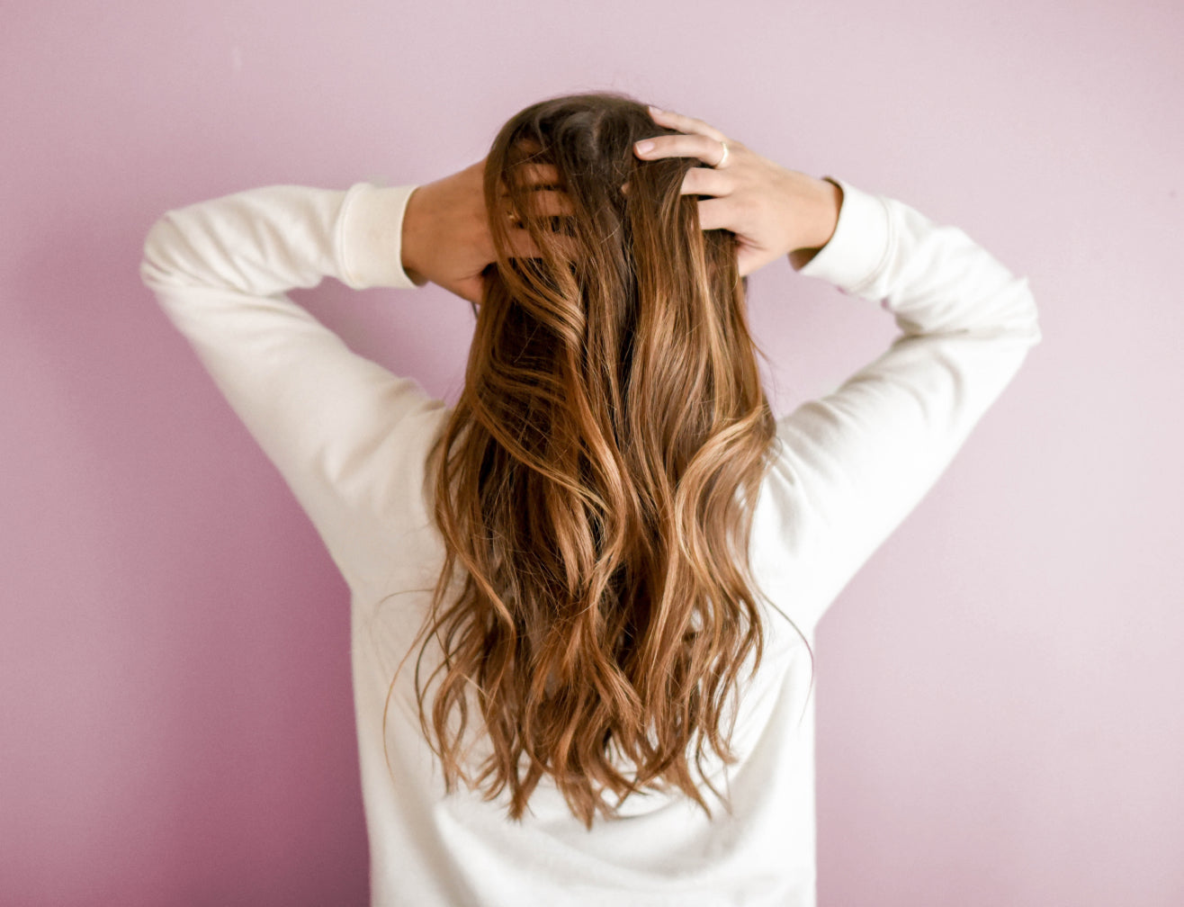 Woman with wavy, long hair faces away, hands on head, against a pink background, highlighting hair health and beauty.