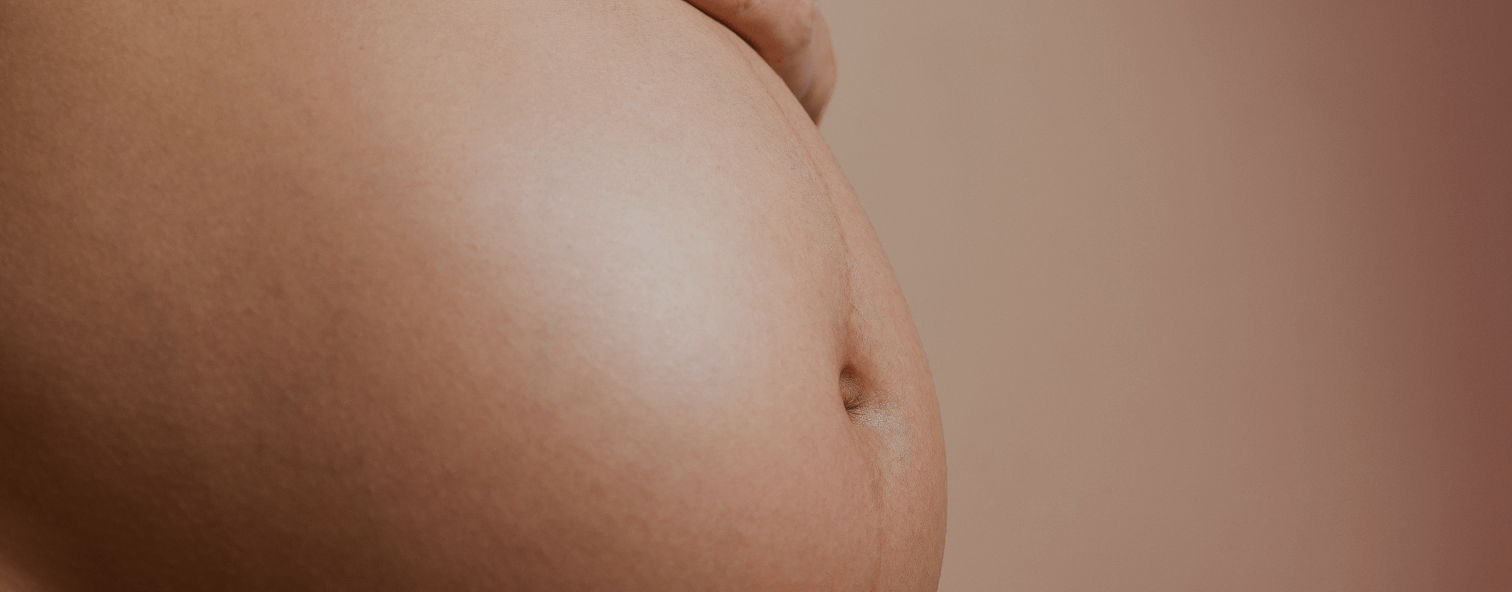 Close-up of a pregnant belly with a hand gently resting on top. The skin is smooth and highlighted against a warm, neutral background.