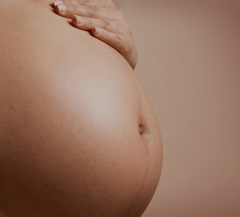Pregnant belly with a hand resting on top, against a soft brown background, highlighting maternal beauty and wellness.