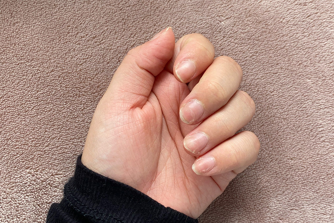 Hand with natural, unpolished nails on a soft pink surface, wearing a black sleeve. Focus on nail health and beauty.