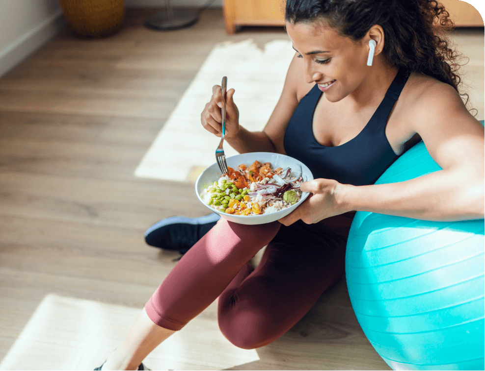 Woman in workout attire eating a healthy salad beside an exercise ball. She wears earbuds and sits relaxed on a wooden floor, smiling.