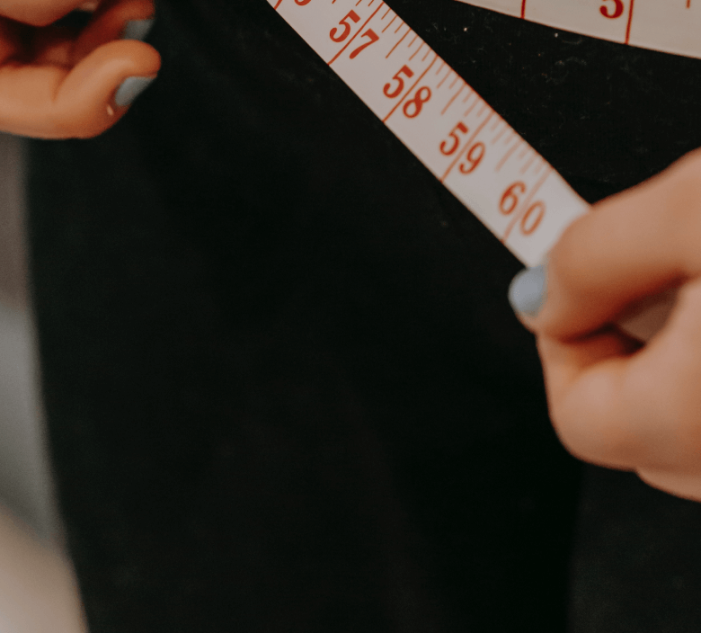 Close-up of hands holding a measuring tape around a waist, indicating a focus on fitness, wellness, and body measurement.