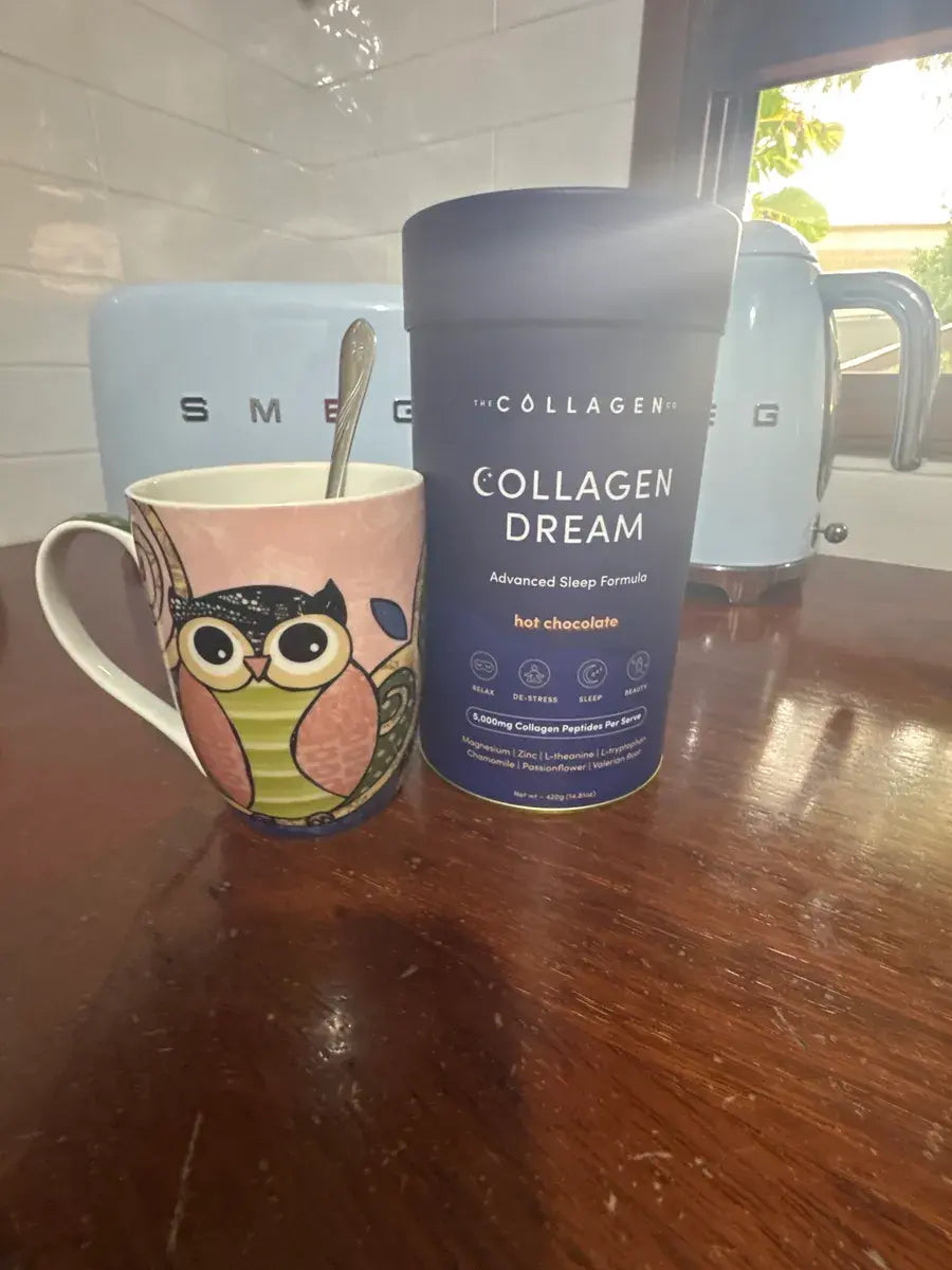 A pink owl-patterned mug with a spoon sits next to a container of Collagen Dream Advanced Sleep Formula hot chocolate on a wooden table, with light blue appliances in the background.