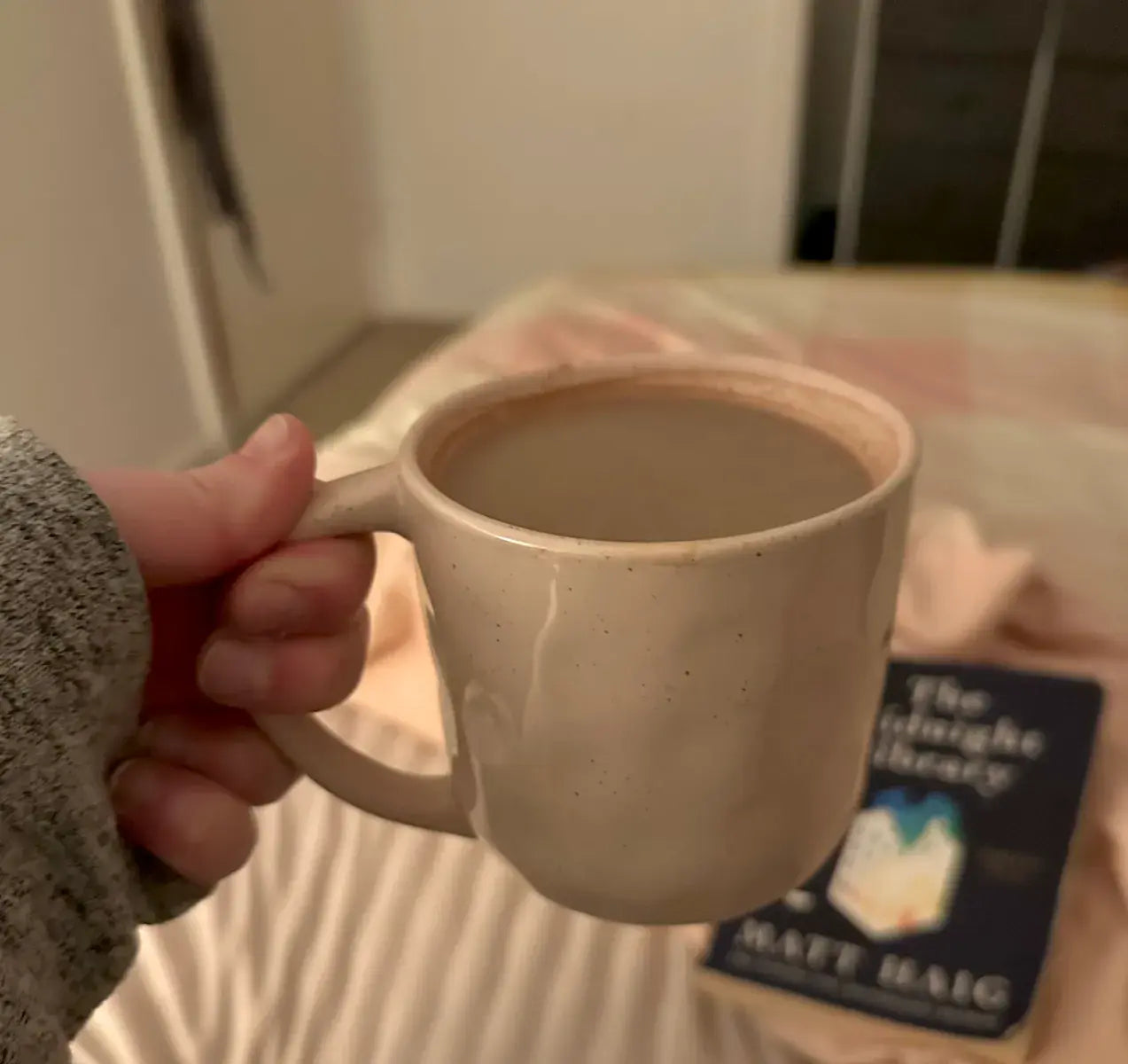 A hand holds a beige mug filled with a hot drink. In the blurry background, a bed with a pinkish blanket and a book titled The Midnight Library by Matt Haig are visible.