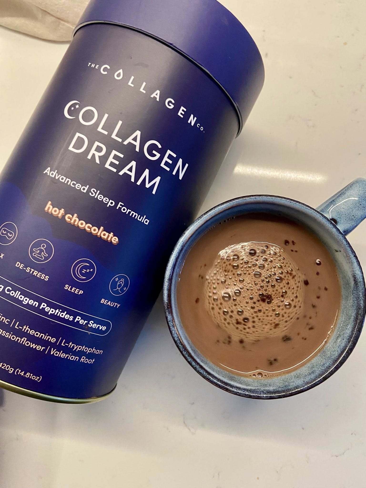 A blue container labeled Collagen Dream Advanced Sleep Formula Hot Chocolate sits next to a blue mug filled with frothy hot chocolate on a white surface.