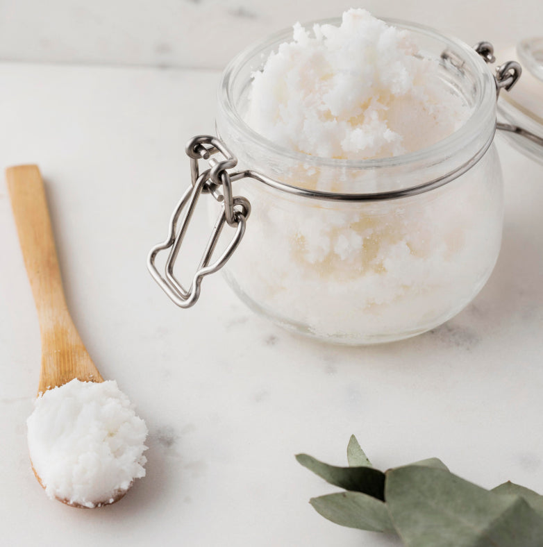 A glass jar filled with white, fluffy body scrub sits open on a white surface next to a wooden spoon holding a scoop of the scrub. Green leaves are partially visible in the corner.