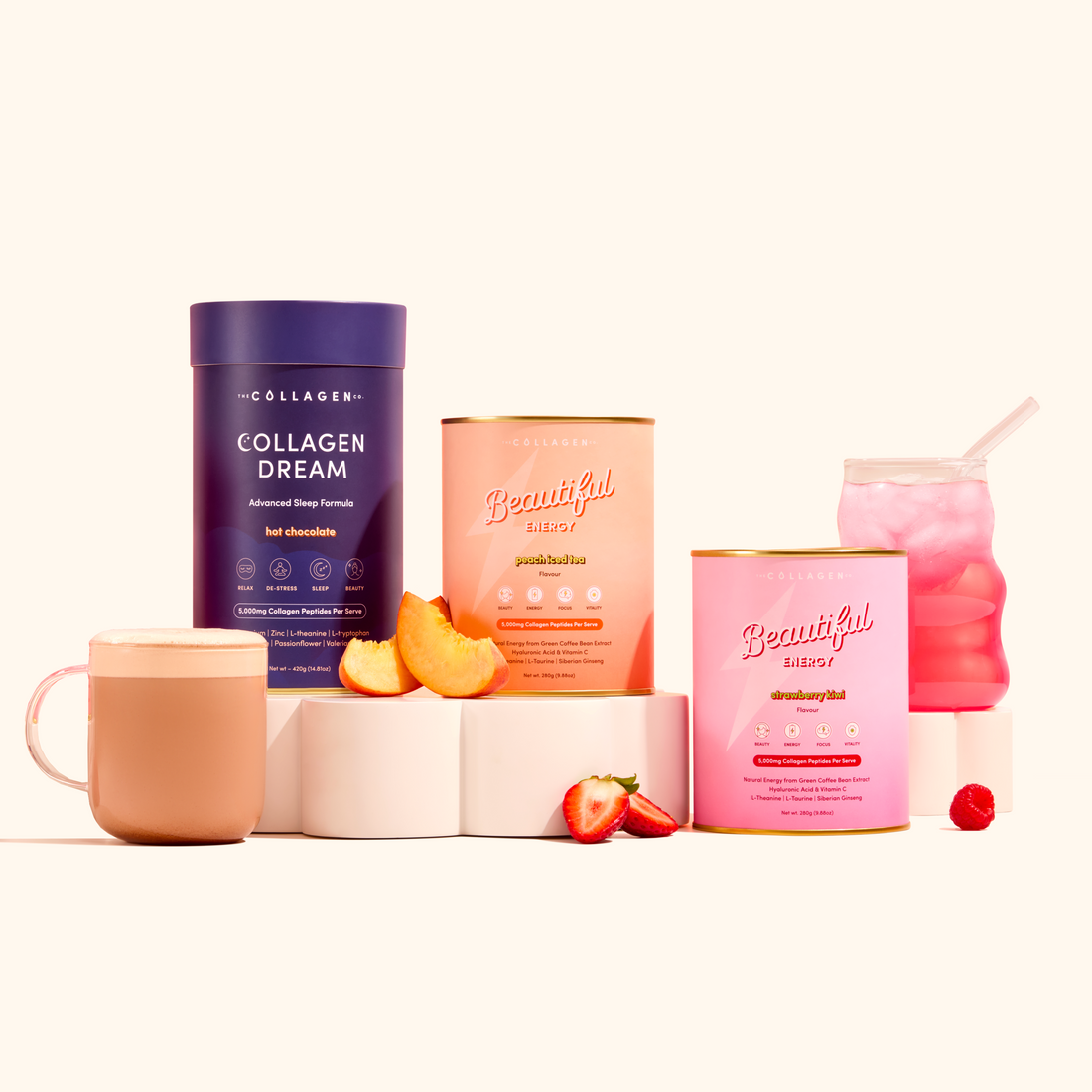 Three colorful Glow AM to PM collagen supplement containers by The Collagen Co. are displayed with a glass mug of Collagen Dream Hot Chocolate, a pink Beautiful Energy drink, peach slices, strawberries, and raspberries on a white surface.
