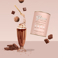 A glass of The Collagen Co. Chocolate Milkshake Collagen Meal Replacement (756g), topped with whipped cream, is surrounded by floating chocolate pieces, a gold spoon with powder, brownies, chocolate bars, and a pink Glow Shake canister. Contains hydrolysed collagen peptides.