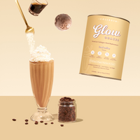A glass of The Collagen Co. Iced Coffee Collagen Meal Replacement (784g) topped with whipped cream, a spoon pouring powder, a scoop of ice cream, the Glow Shake canister, and coffee beans are displayed on a beige background.