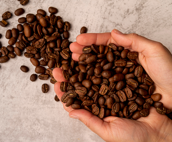 A pair of hands holds a pile of roasted coffee beans, with more coffee beans scattered on a light, textured surface in the background.