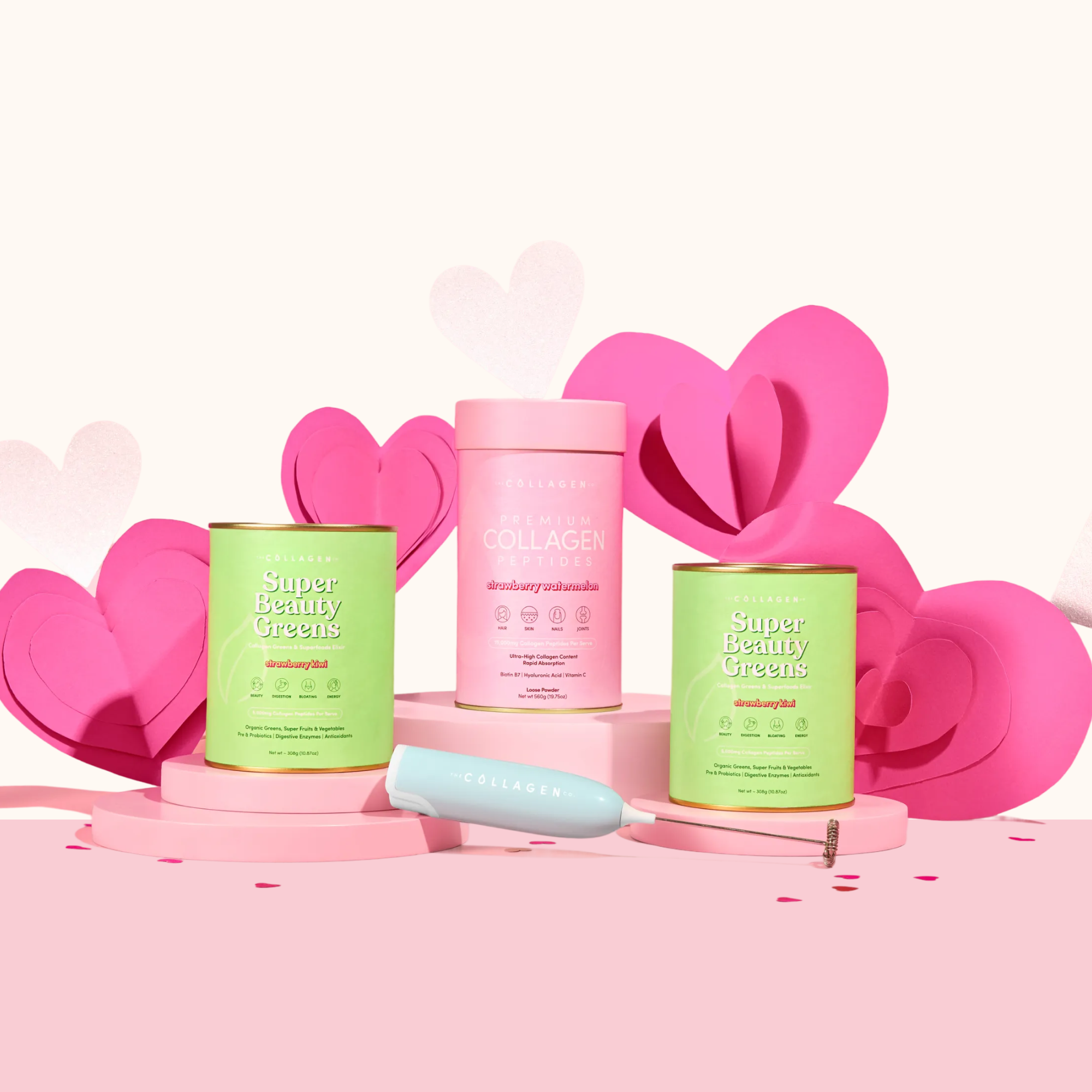 The Collagen Co.'s Gut Feelings Beauty Bundle—two green and one pink canisters—sits on a pink surface with heart decorations, plus a white collagen spoon and whisk in front.