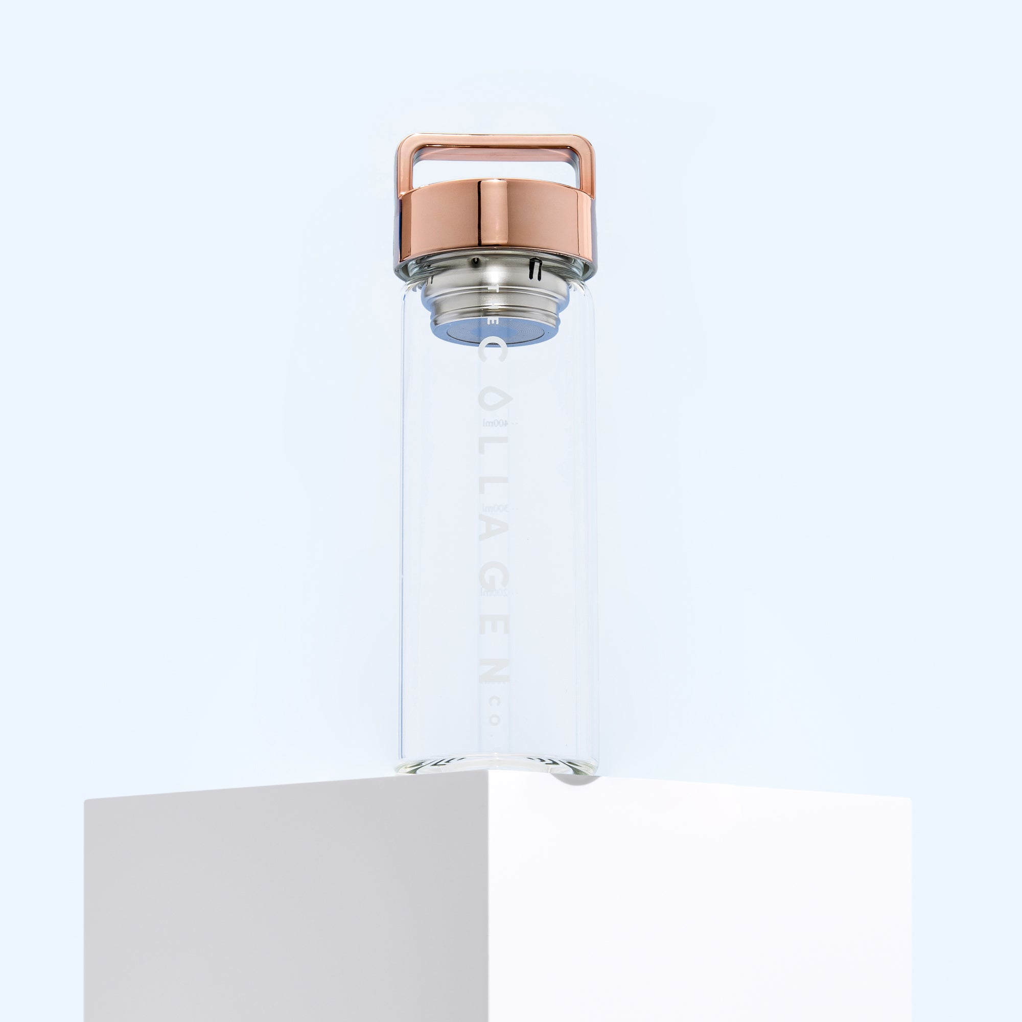 A clear Infuser - 500ml by thecollagen.co sits on a white pedestal with a rose gold cap and handle against a light blue background. "COLLAGEN" is vertically printed on the bottle, showcasing its function as a stylish collagen infuser.