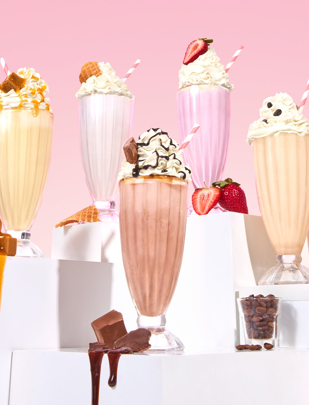 Five colorful milkshakes in tall glasses topped with whipped cream, fruit, chocolate, and waffle cones are displayed on white platforms against a pink background. Chocolate pieces, coffee beans, and strawberries are scattered nearby.