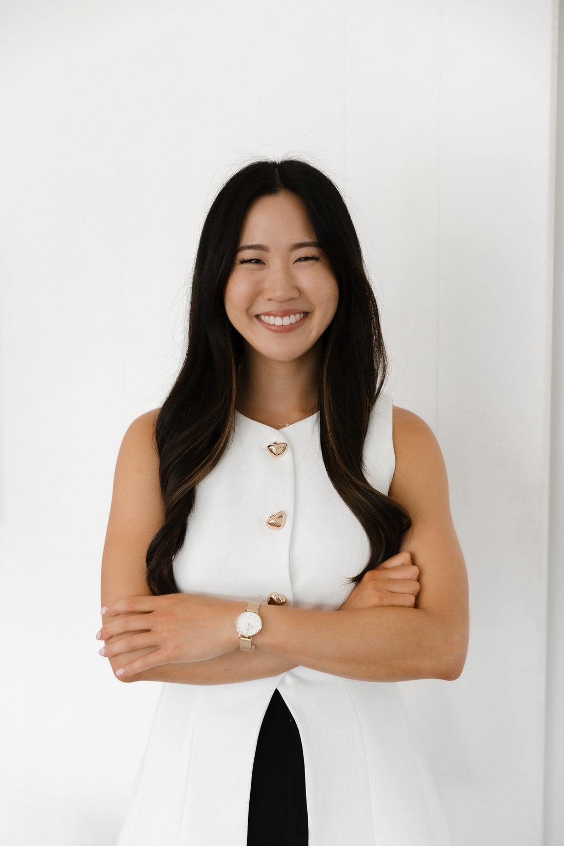 A woman with long dark hair, wearing a sleeveless white top, stands smiling with her arms crossed in front of a plain white background.