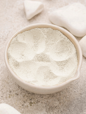 A bowl filled with fine white powder, showing several smooth, rounded indentations on the surface. White stones are placed around the bowl on a light, textured background.