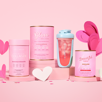 The Collagen Co. Love Your Glow Bundle, featuring four collagen supplement containers and a clear shaker with a pink drink, is displayed on pink platforms with paper hearts against a pastel pink backdrop for a beautiful energy boost.