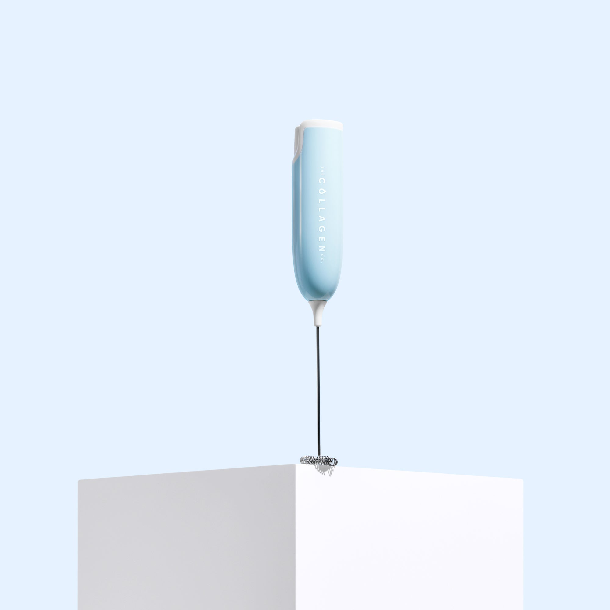 The Collagen Co USA Collagen Mixer, a light blue portable handheld device with a white top, stands upright on a white cube against a pale blue background.