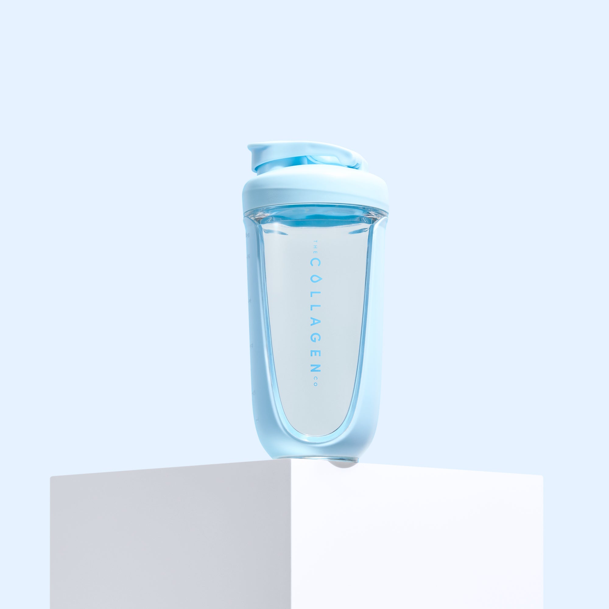 The Collagen Co. Collagen Protein Shaker - 600ml, featuring a transparent body with light blue lid and base, labeled "THE COLLAGEN," is displayed on a white pedestal against a pale blue background.