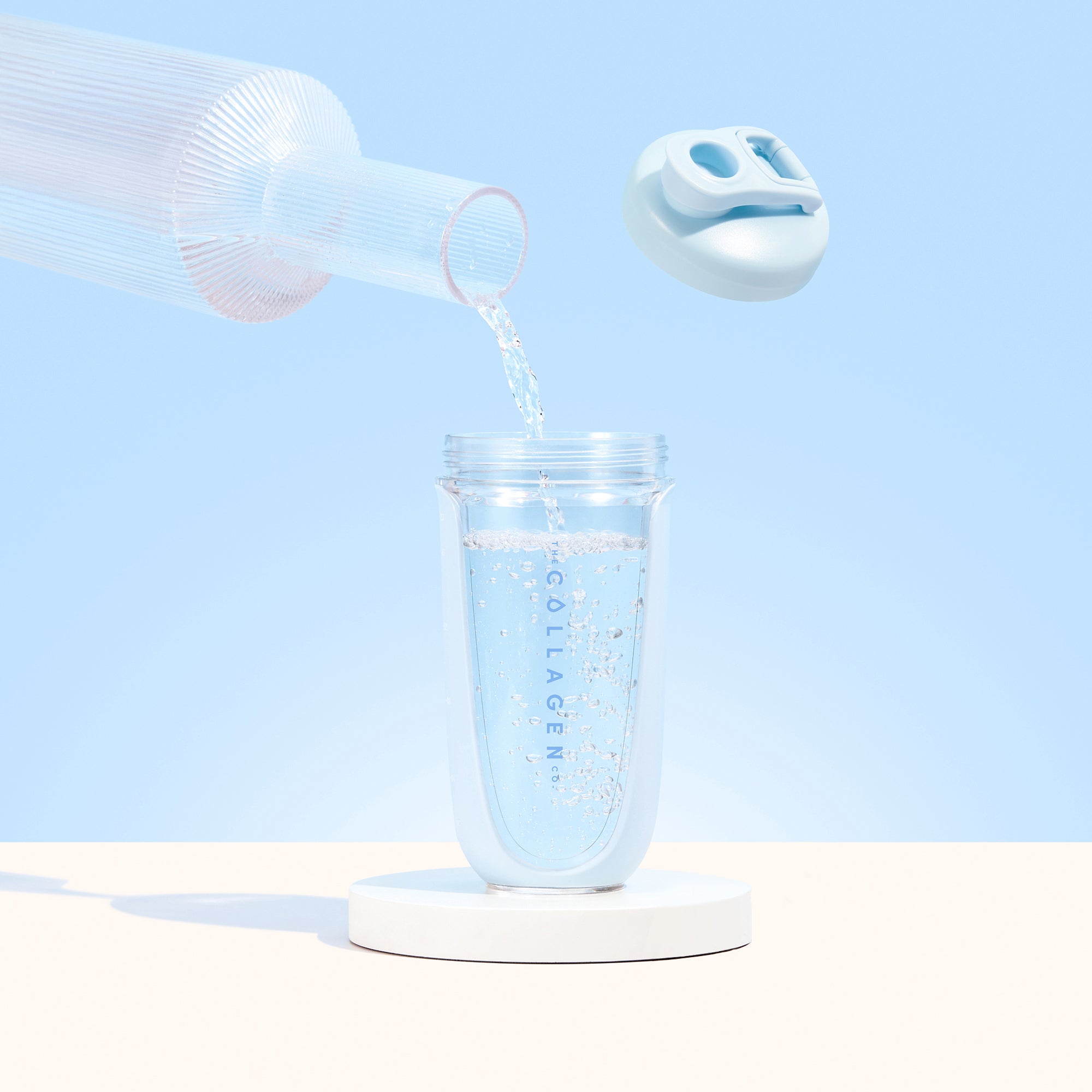 A clear pitcher pours water into The Collagen Co. Collagen Protein Shaker - 600ml, labeled COLLAGEN with a light blue lid above, set against a pastel blue background.