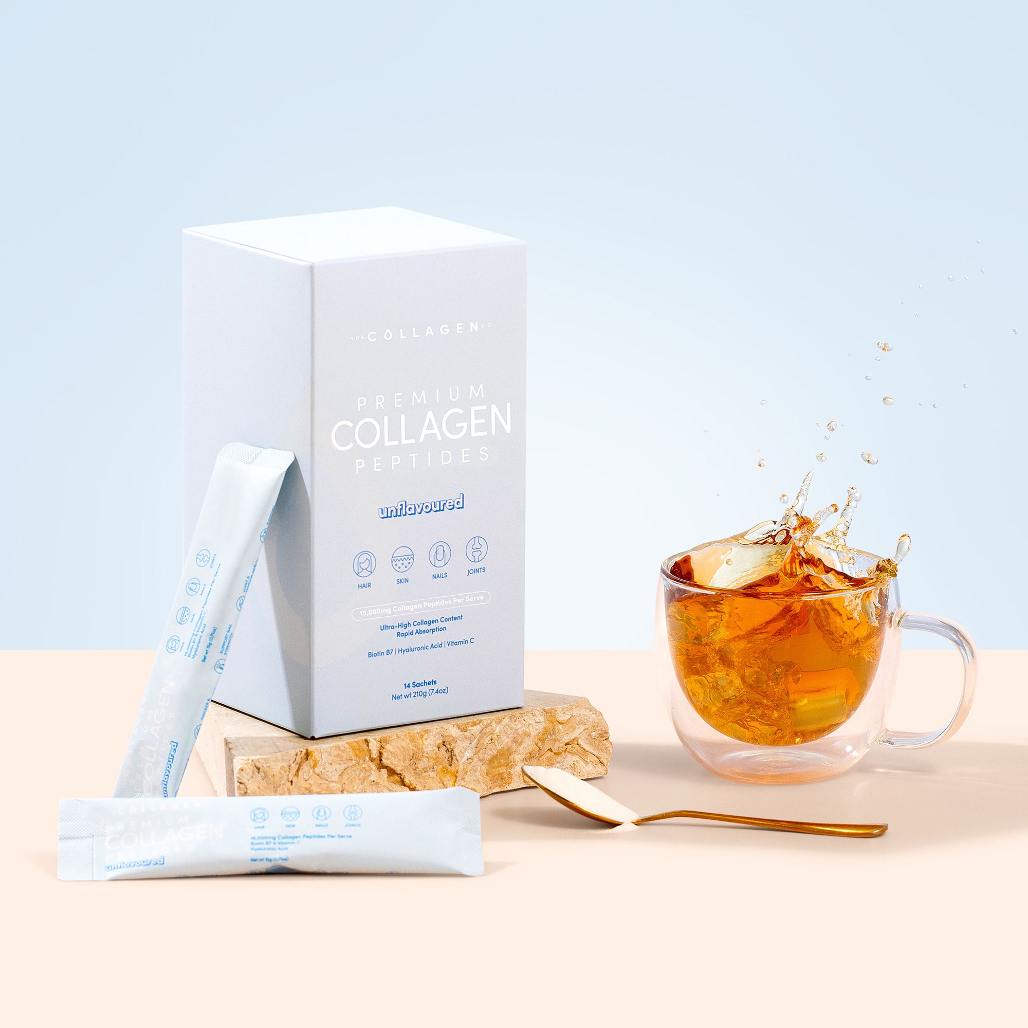A box of thecollagen.co Unflavored Collagen Sachets (210g) is displayed with individual sachets, a gold spoon, and a glass cup with amber liquid—ideal for healthy hair, skin, and nails on a bright background.