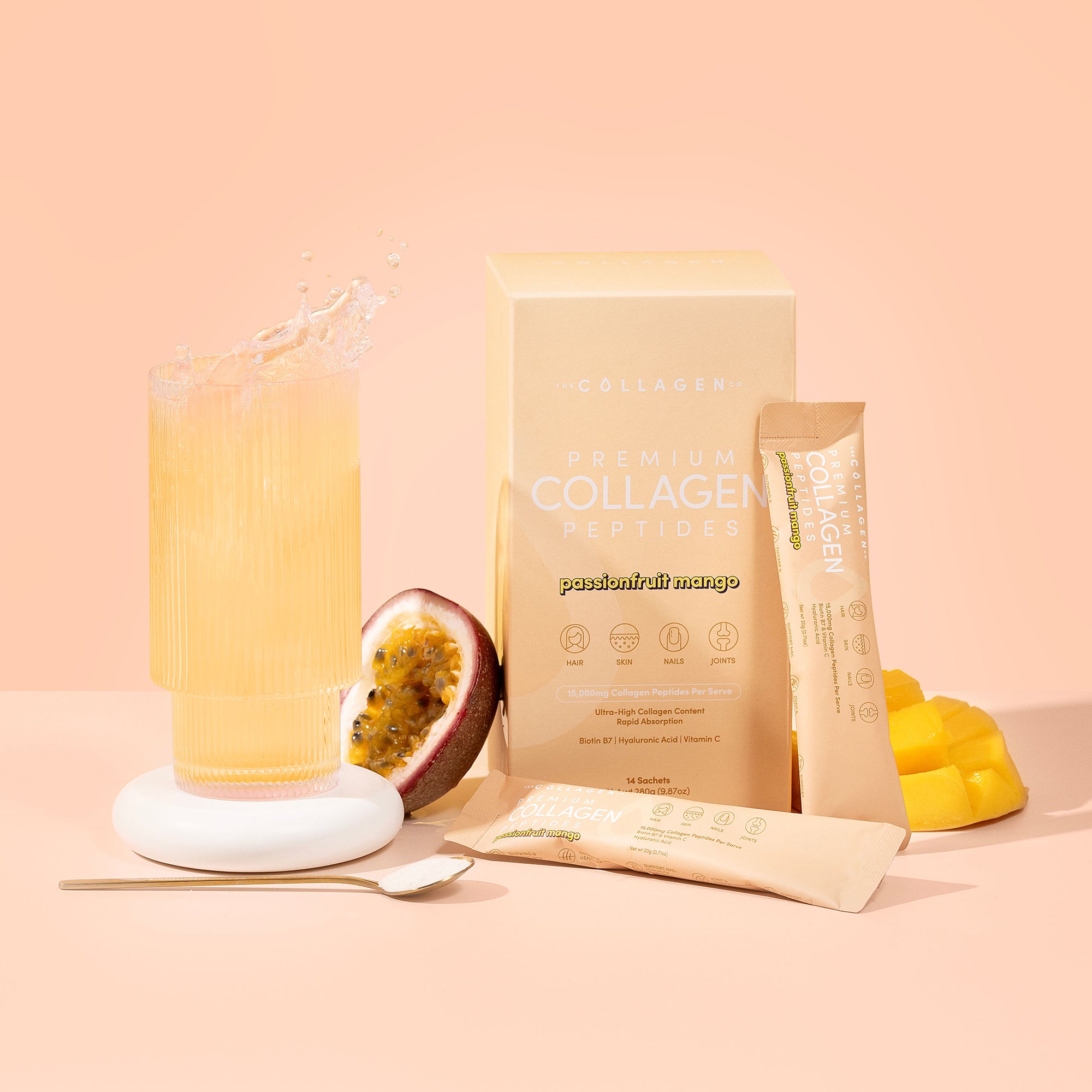 A vibrant glass of yellow drink splashes beside passionfruit, mango pieces, spoon, box, and sachets of thecollagen.co’s Passionfruit Mango Collagen Sachets—showcasing Beauty and Wellness with Hydrolysed Bovine Collagen Powder.