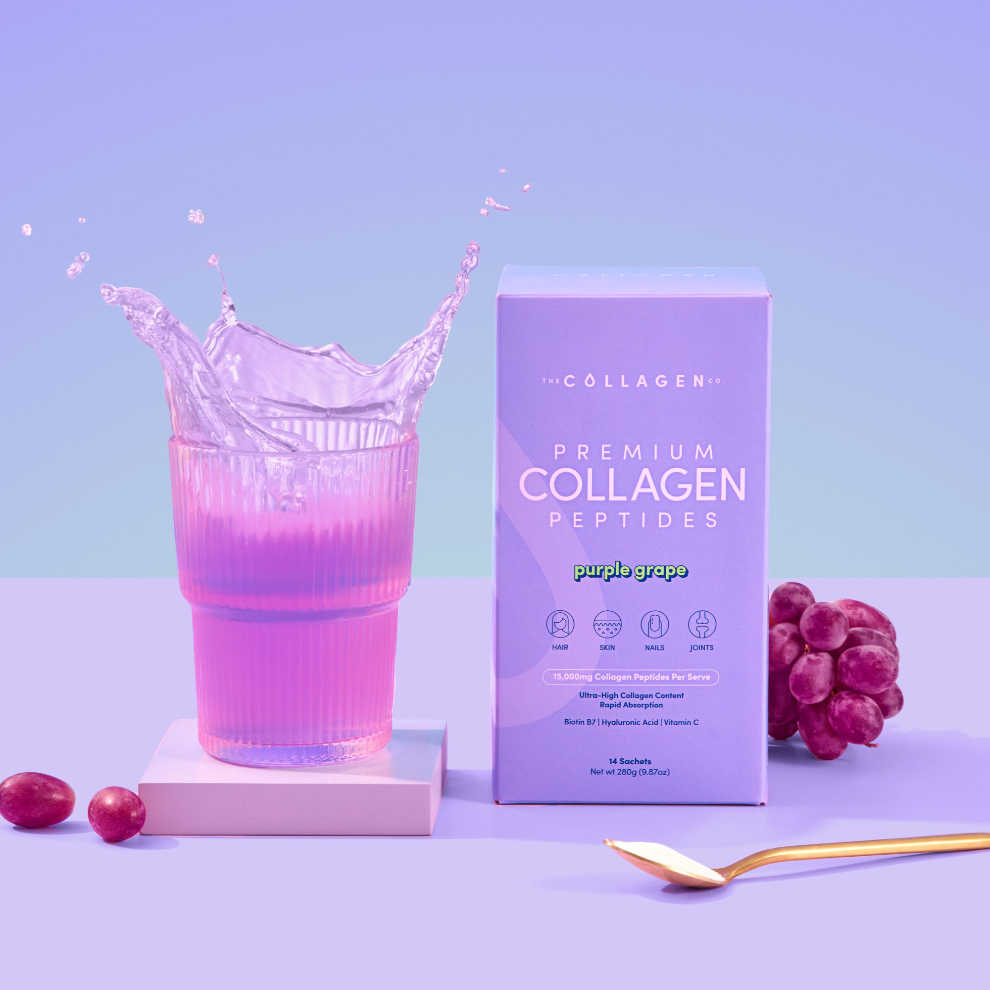 A glass of purple liquid splashes beside a box of The Collagen Co USA Purple Grape Collagen Sachets (280g), with grapes, a gold spoon, and hydrolysed bovine collagen powder on a pastel purple background.