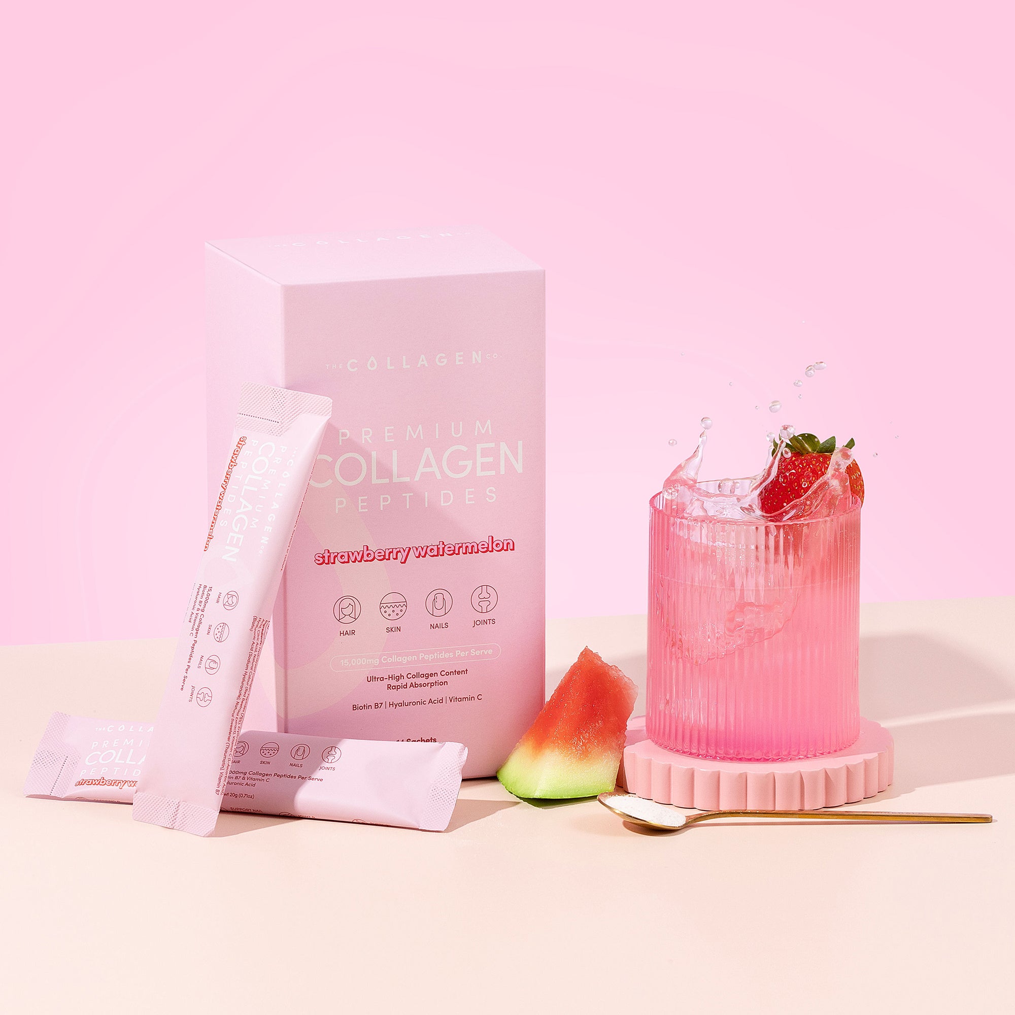 A box of Strawberry Watermelon Collagen Sachets by thecollagen.co stands next to hydrolysed bovine collagen powder sticks, a glass of pink strawberry drink with ice, a watermelon slice, and a gold spoon on a vibrant pink background.