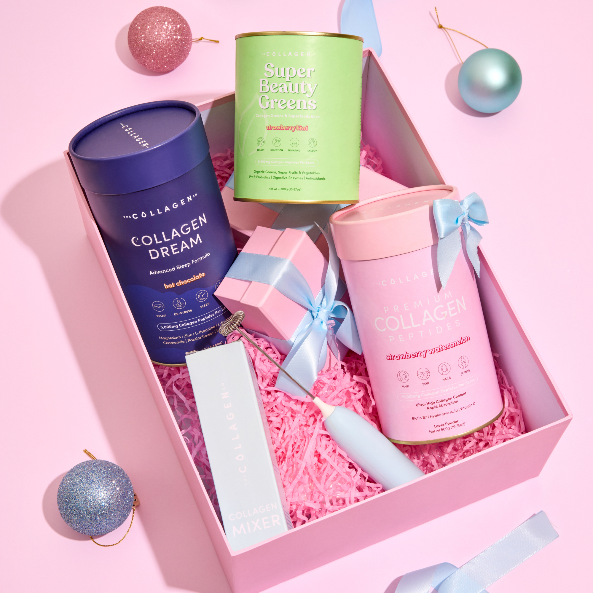 A pink gift box from The Collagen Co. featuring the Reset & Renew Trio—Collagen Dream, Super Beauty Greens, Collagen Peptides—a Collagen Mixer, wrapped gift, and festive ornaments; perfect for gifting or enjoying as a sleep hot chocolate treat.