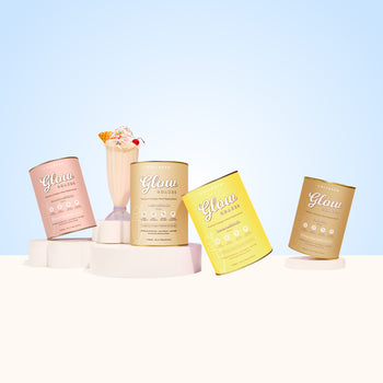 The Collagen Co.’s Shake & Glow Bundle features four vibrant protein powder containers and a whipped cream-topped meal replacement shake, all beautifully arranged on white platforms against a light blue backdrop.