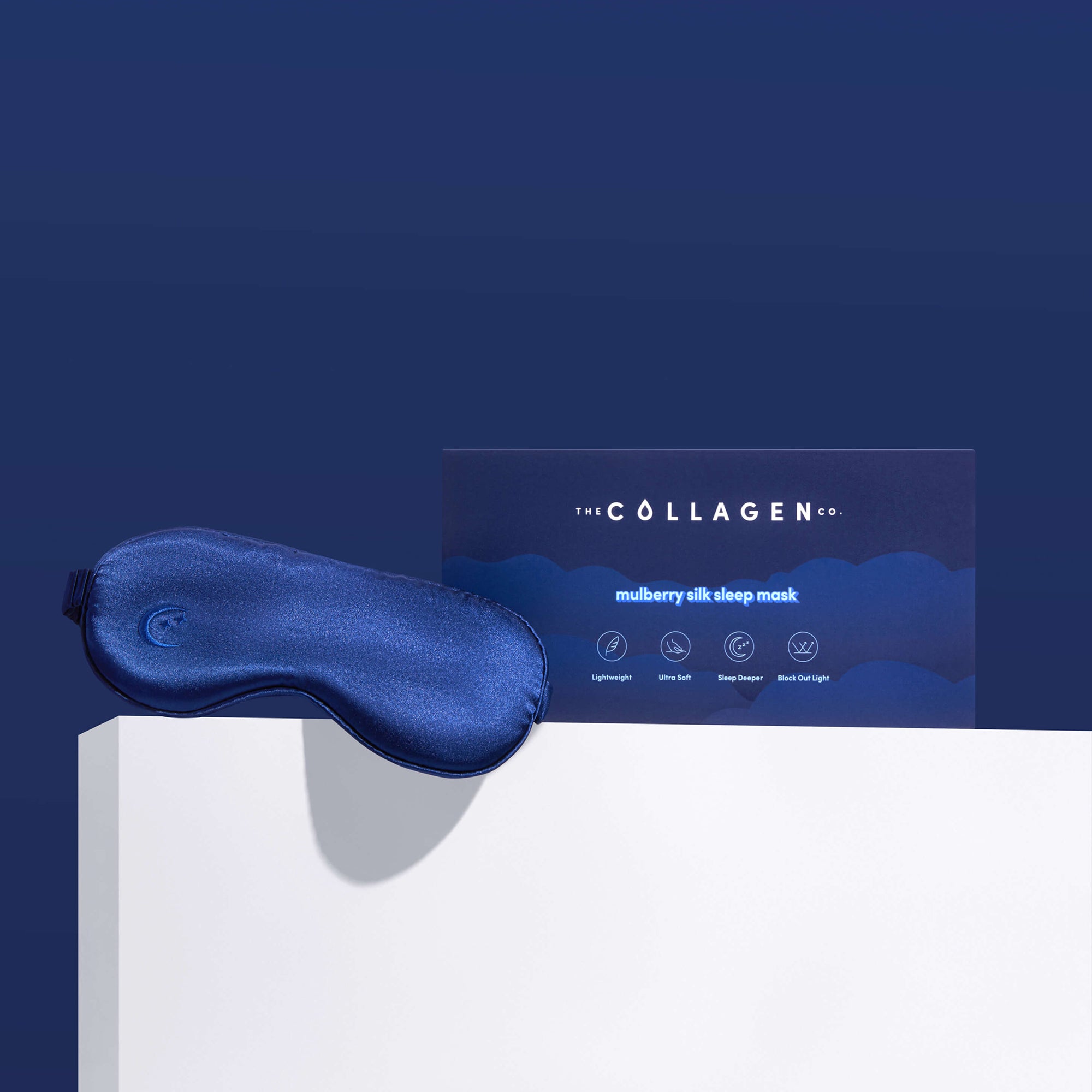 A Mulberry Silk Sleep Mask by The Collagen Co. in navy blue sits on a white surface beside its branded box, all set against a dark blue background.