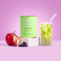 A green canister of The Collagen Co USA’s Apple Blackcurrant Super Beauty Greens (308g) sits with a red apple, apple slices, blackcurrants, and a glass of green drink with a straw on a pink background.