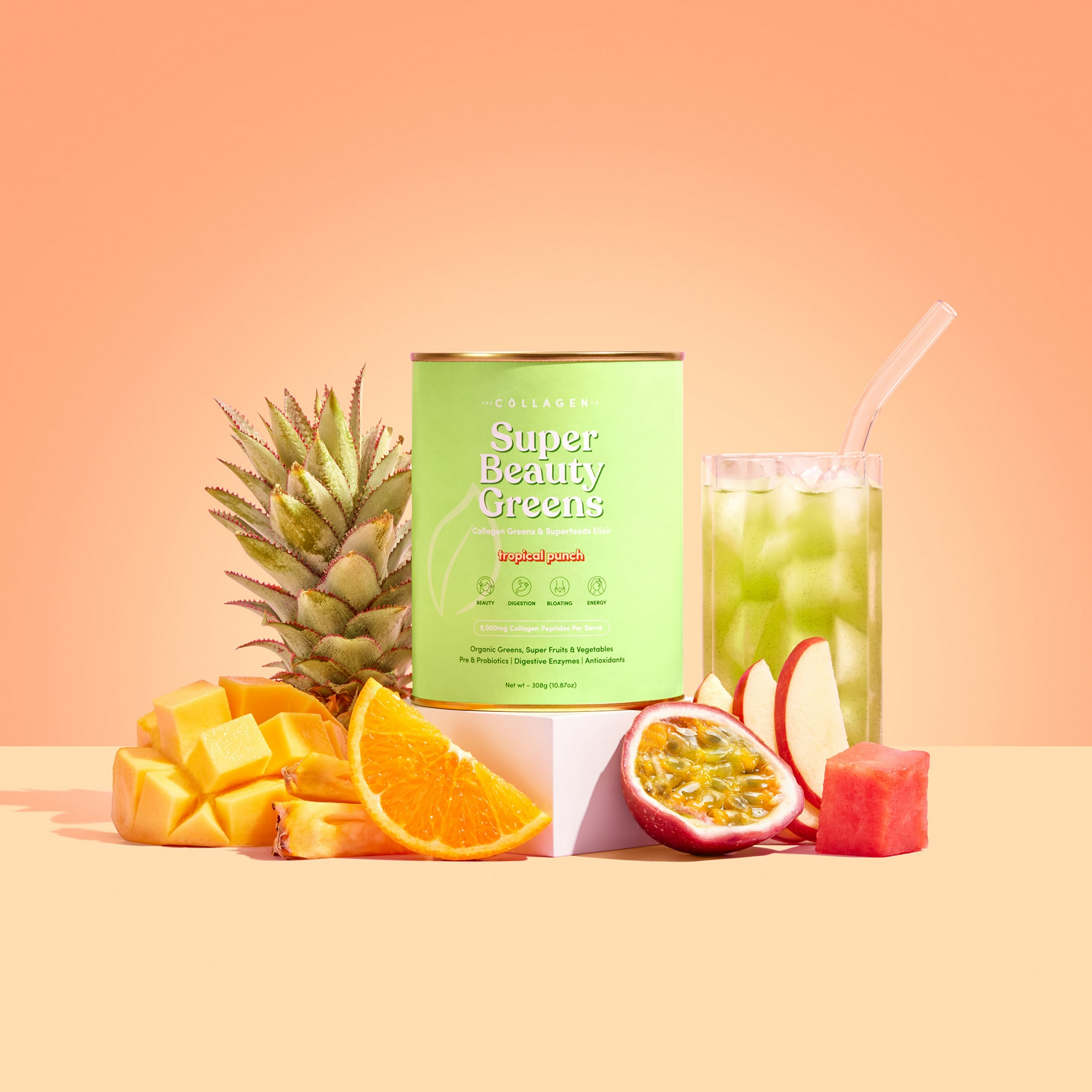 A canister of The Collagen Co USA Tropical Punch Super Beauty Greens - 308g sits beside a glass of green juice, surrounded by pineapple, mango, orange, apple, watermelon, and passionfruit against a peach background.