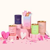 Five colorful containers of The Collagen Co.’s The Complete Glow Affair Bundle are arranged on pink platforms with paper hearts in pink, red, and white—featuring collagen peptides for beauty and gut health in a vibrant, feminine display.