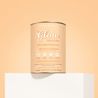 A cylindrical tub of The Collagen Co. Salted Caramel Milkshake Collagen Meal Replacement (756g) rests on a white pedestal against a peach backdrop, featuring peach packaging with white and gold text.